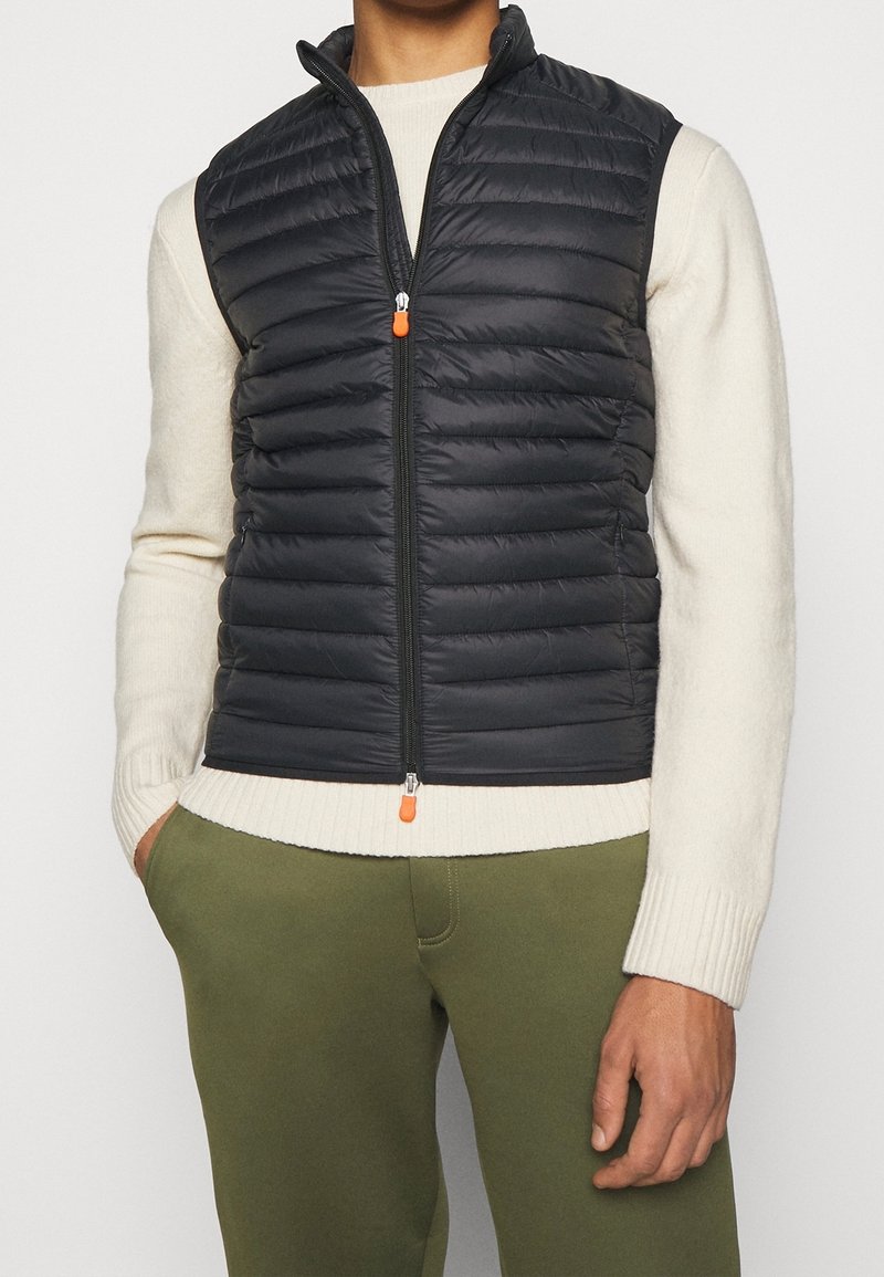 Homme portant un gilet matelassé noir sans manches sur un pull crème et un pantalon vert olive, debout avec une main dans la poche.
