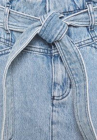 Ljusblå denimtyg med en knuten bälte-detalj. Texturen är tydligt vävd, med vit sömnad längs sömmarna och kanterna.