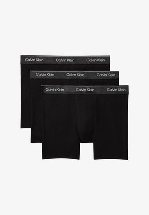 Muskulöser Mann steht barfuß vor einfachem Hintergrund, trägt schwarze Boxershorts und richtet mit beiden Händen den Hosenbund.