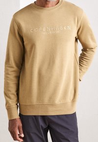 Sweatshirt beige à col rond, brodé "COPENHAGEN LINDBERGH" sur le devant, texture lisse, poignets et ourlet côtelés, coupe décontractée.