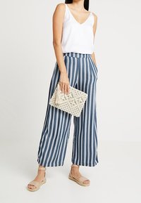 Obcisła biała koszulka bez rękawów, granatowe i białe spodnie w szerokim kroju w paski, beżowe espadryle oraz pleciona torba kopertówka z detalem w postaci frędzla.