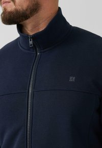 Marine fleece jack met een hoge kraag, volledige rits aan de voorkant en een logo-accent. Heeft een gladde textuur en contrasterende stiksels.
