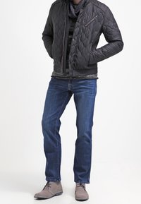 Vadderad svart bomberjacka med en dragkedjeficka, lagd över en mörkrandig skjorta, ihop med blåjeans och grå snörskor.