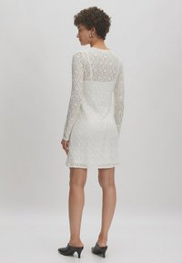 Weißes Spitzenkleid mit langen Ärmeln, strukturiertem Blumenmuster, tailliertem Oberteil und ausgestellt geschnittenem Saum. Das Model kombiniert es mit schwarzen Mules.