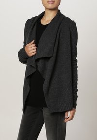Ciemnoszary, oversize kardigan z miękkiego, teksturowanego materiału. Ma zwiewny przód i prążkowane rękawy, noszony na czarnym topie.