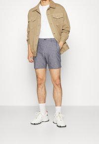 Hnědá bunda s knoflíkovými kapsami, bílý tričko, šedé šortky a bílé tenisky. Ponožky jsou bílé s červenými akcenty. Jednoduchý design a casual střih.