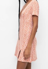 Vestido de manga corta en naranja con estampado floral blanco, diseño de escote en V, largo hasta la rodilla y con aberturas laterales para facilitar el movimiento. Textura de tela suave.