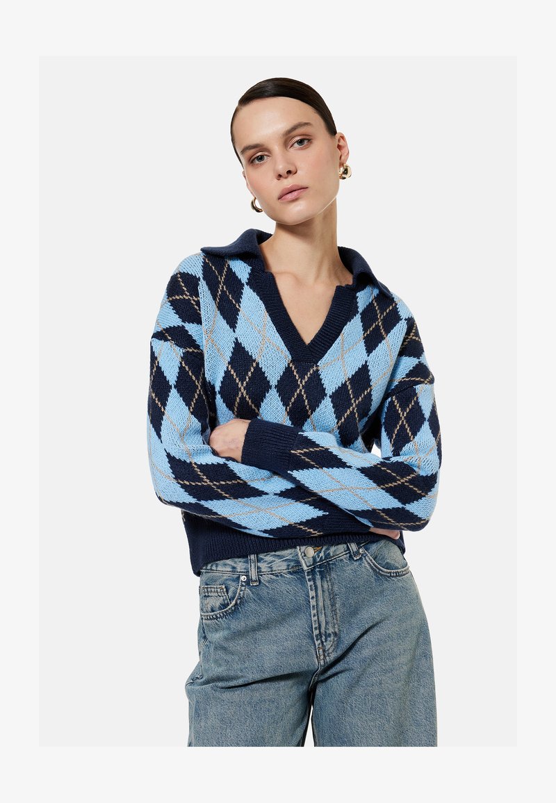 Vrouw draagt een blauw en marineblauw argyle trui met V-hals en kraag, gecombineerd met lichtblauwe spijkerbroek, poseert met gekruiste armen.