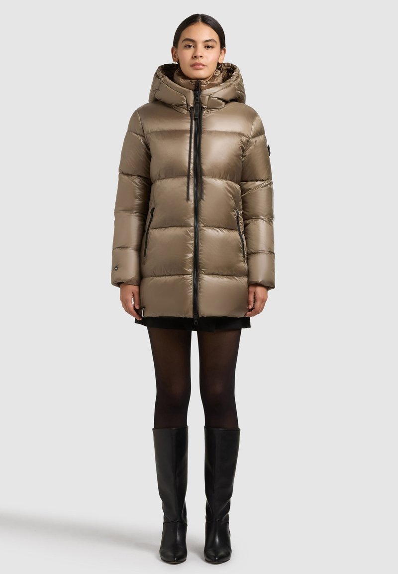 Prošívaná bunda světle hnědé barvy s kapucí, vertikálním zipem, bočními kapsami a prošívanou texturou. Nositelná přes krátké černé šaty a punčochy.