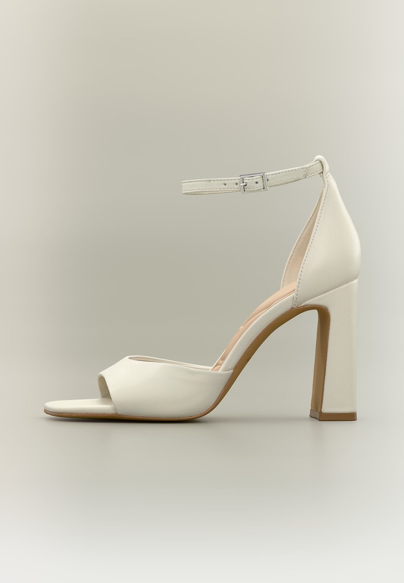 Sandale blanche à talons hauts avec bride cheville, bout ouvert, semelle intérieure rembourrée et talon bloc sur fond neutre.