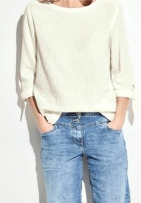 Blusa bianca con texture e maniche a tre quarti, abbinata a jeans in denim azzurro chiaro con chiusura a bottoni e tasche laterali.