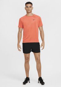 Ung mann står og vender ansiktet fremover iført en oransje Nike T-skjorte, svarte shorts og svarte Nike sneakers på hvit bakgrunn.