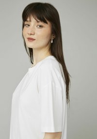 T-shirt blanc à manches courtes avec une coupe décontractée. Encolure ronde, texture lisse et design simple. Le modèle porte des boucles d'oreilles en perles.