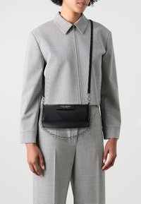 Black leather crossbody bag with a chain strap, featuring a smooth finish and a rectangular shape. Branded tag in white on the front.