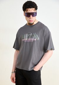 Pelēka īsām piedurknēm t-kreklu, kas rotāts ar atstarotāju "ALPHA STUDIO EDITION" logotipu purpura un rozā krāsā, saskaņojumā ar tumšiem biksēm un pārmērīgi lielām purpura saulesbrillēm.