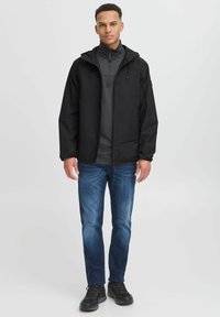 Zwarte softshell jas met een rits aan de voorkant en een capuchon, gedragen over een grijze top. Gepaard met blauwe jeans en zwarte sportschoenen.