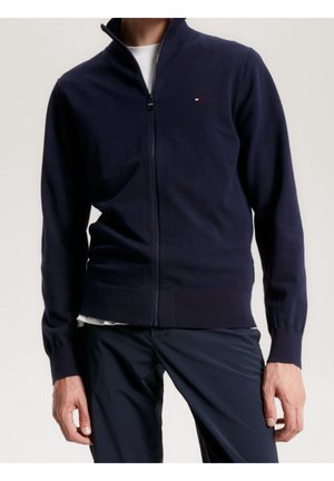 Homme portant un pull zippé bleu marine avec un petit logo rouge, blanc et bleu sur la poitrine, associé à un pantalon foncé et un t-shirt blanc en-dessous.