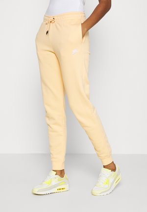 Henkilö, jolla on vaaleankeltaiset Nike-housut ja niihin sopivat lenkkarit, seisoo käsillä taskuissa yksiväristä taustaa vasten.