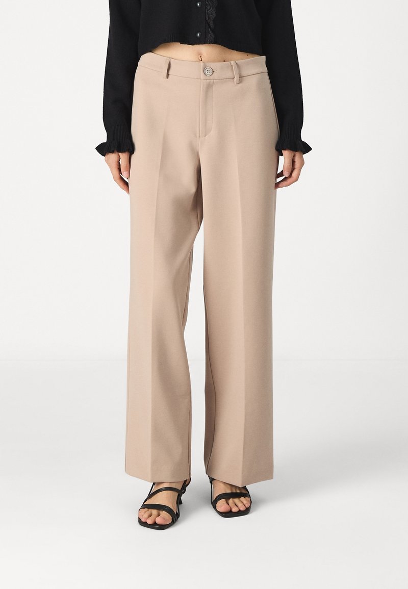 Pantalon beige taille haute à jambes larges porté avec des sandales noires à lanières et un top court noir à manches longues avec bordure en dentelle.