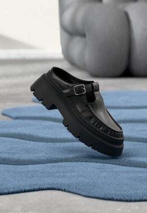 Sort læderloafer med chunky profil sål og sølvspænde, placeret i en vinkel på et struktureret blåt tæppe.