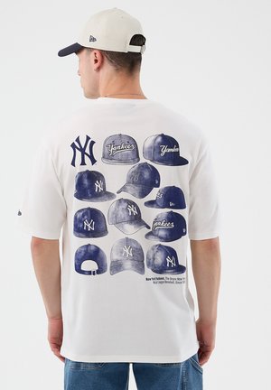 Uomo che indossa una maglietta bianca con multiple illustrazioni di cappellini da baseball New York Yankees blu sulla schiena, abbinata a jeans blu e cappellino beige.