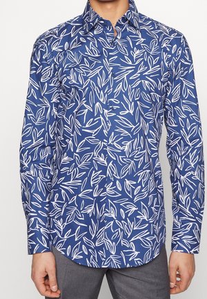 Niebieska koszula na długi rękaw z białymi, abstrakcyjnymi wzorami liści, noszona z szarymi spodniami, widoczna górna część ciała od szyi do bioder.