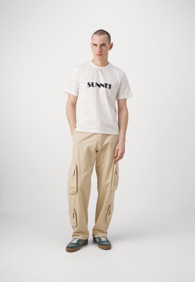 Tricou alb din bumbac cu "SUNNEI" imprimat în negru, pantaloni cargo bej cu buzunare laterale și adidași verzi și gri cu șireturi deschise.