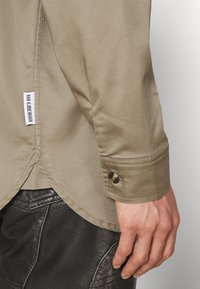 Main droite à côté du poignet d'une chemise beige à manches longues avec bouton et étiquette, sur un pantalon en cuir noir texturé.