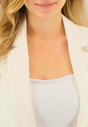 Collier en or avec une chaîne lisse et fine, exposé sur un haut blanc et sous un blazer blanc. Le collier présente un design simple et élégant.