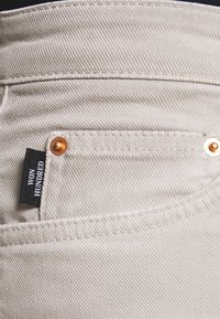 Pantalones de mezclilla beige con tejido texturizado, que presentan una pequeña etiqueta negra "WON HUNDRED" y detalles de botones de color cobre en el bolsillo.