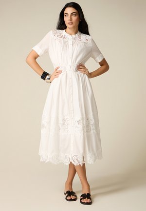 Donna con lunghi capelli scuri che indossa un vestito midi bianco in pizzo con maniche corte, con bottoni davanti e sandali neri, in piedi con le mani sui fianchi.