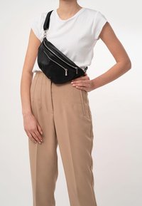 Borsa a tracolla nera in pelle con tre scomparti con cerniera, caratterizzata da una superficie texturizzata e una tracolla regolabile, indossata sopra pantaloni beige a vita alta.