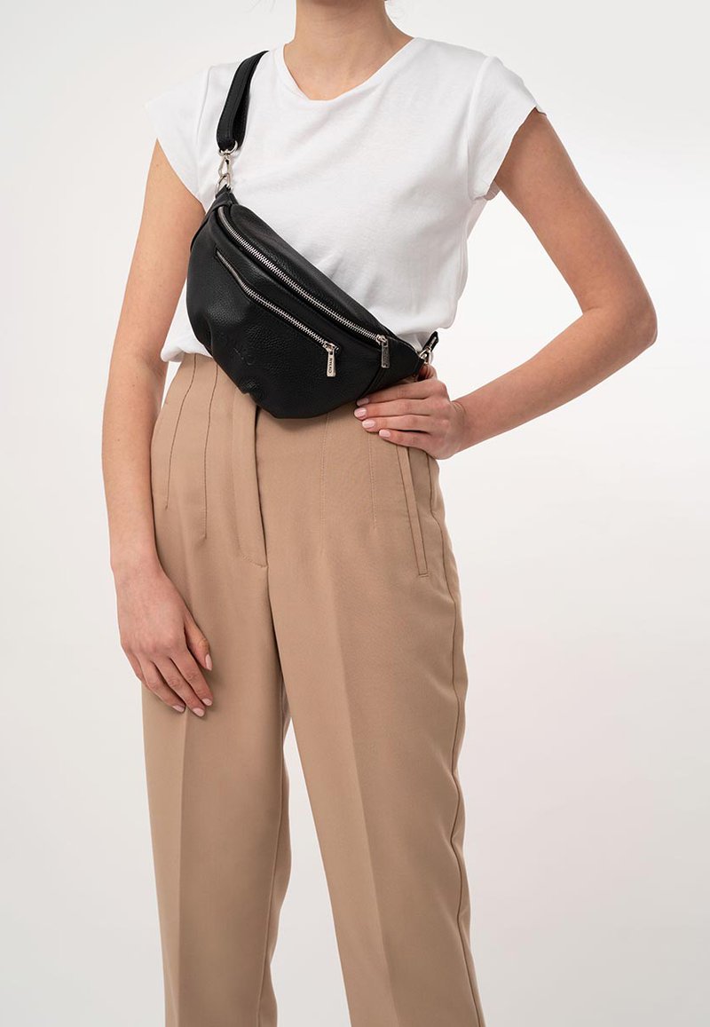 Borsa a tracolla nera in pelle con tre scomparti con cerniera, caratterizzata da una superficie texturizzata e una tracolla regolabile, indossata sopra pantaloni beige a vita alta.