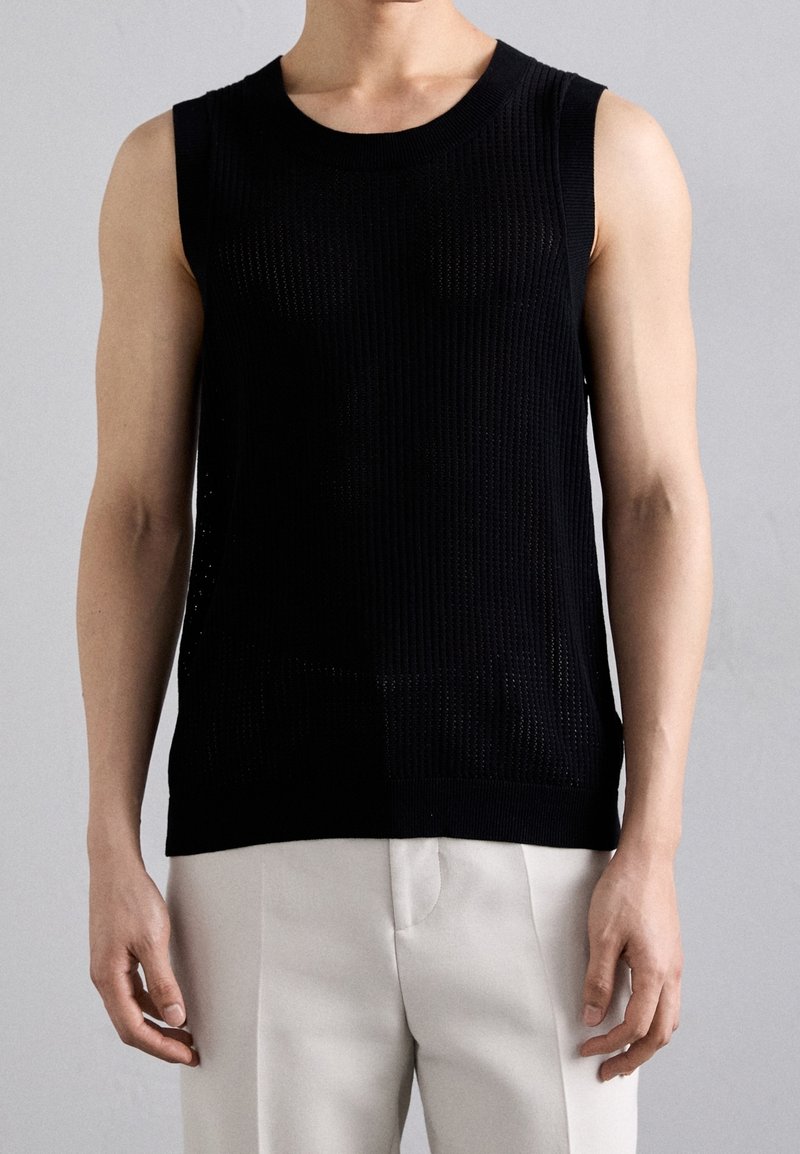 Maglione nero senza maniche in maglia con texture a coste, caratterizzato da un collo rotondo e delicate perforazioni, abbinato a pantaloni di colore chiaro.