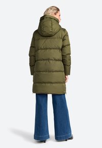 Lange olijfgroene pufferjas met capuchon, met verticale stiksels en zijritsen. Gecombineerd met wijde blauwe denim broek.