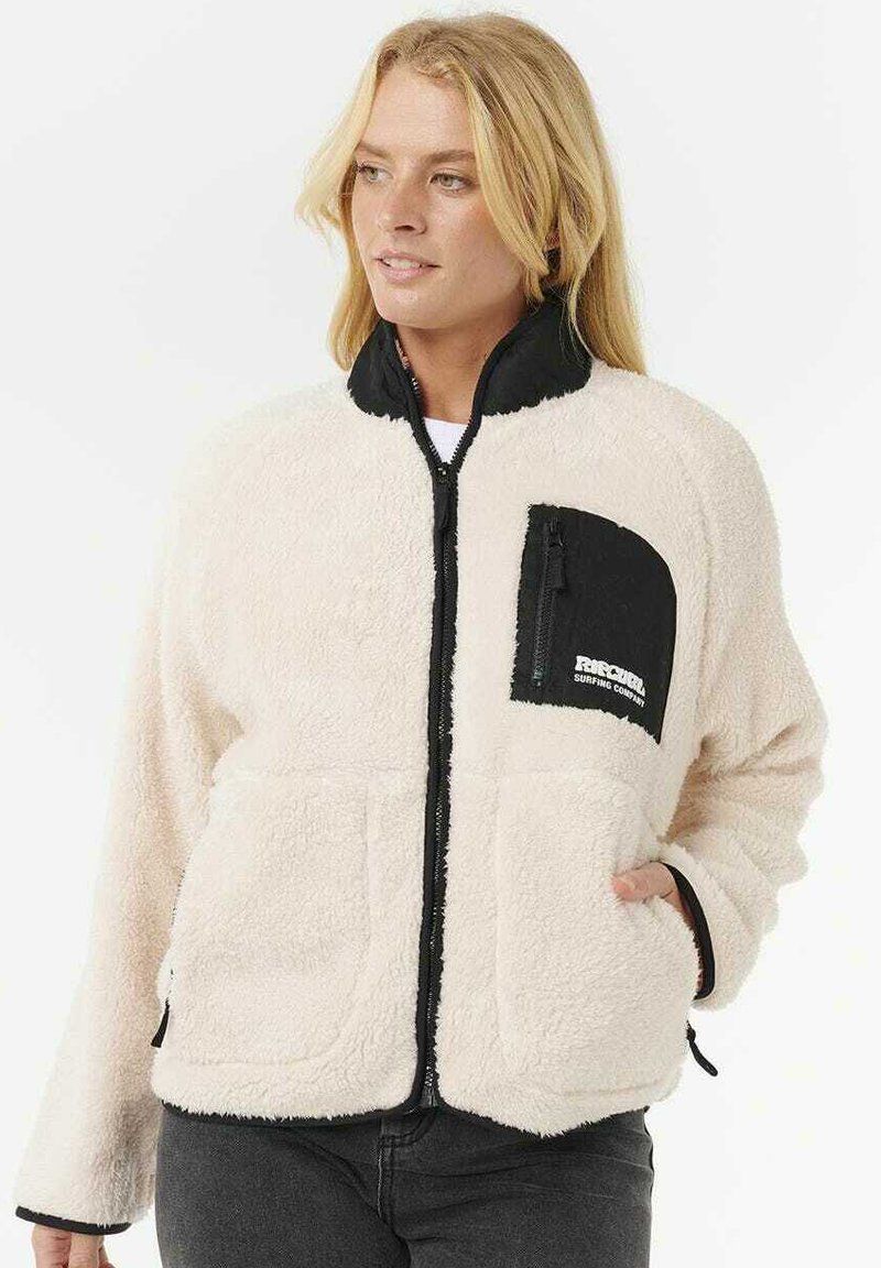 Blonde vrouw in een wit fleece jack met zwarte accenten en een ritssluiting op de borst, staand met één hand in haar zijzak.