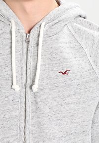 Grå zip-up hoodie med en vit snörning och en liten röd måslogotyp på vänster bröst, som bärs av en person.