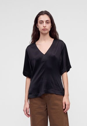 Femme aux longs cheveux foncés portant un haut noir ample à manches courtes en V et un pantalon marron, debout devant un fond clair uni.