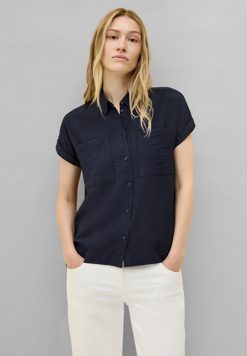 Camicia azzurra a maniche corte con bottoni, caratterizzata da due tasche sul petto, texture liscia e vestibilità rilassata, abbinata a pantaloni bianchi.