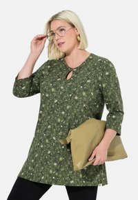 Tumika tumejaa ya kijani kibichi yenye uchapishaji wa maua, shingo yenye mfuniko wa ufunguo, mikono ya 3/4, ikishirikishwa na begi laini la beige. Leggings za mblack zinakamilisha mavazi.