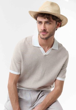 Junger Mann mit lockigem Haar, der ein beiges gestricktes Poloshirt, hellgraue Hose und einen hellen Strohhut trägt, schaut leicht zur Seite.