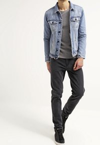 Ljusblå denimjacka med slitna detaljer och två bröstfickor, kombinerad med en grå T-shirt och svarta slim-fit jeans. Svarta sneakers.