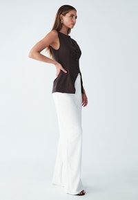 Hnědá bezrukávová vesta s knoflíky, spárovaná s bílými kalhotami širokého střihu. Hladká textura, minimalistický design a boční rozparky pro snadný pohyb.