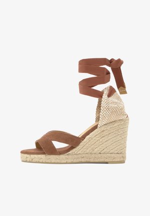 Sandalia alpargata de cuña con plataforma trenzada de yute, tiras de tela marrón cruzadas sobre los dedos y largo lazo para atar en el tobillo con contrafuerte de talón tejido.