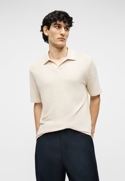 Jeune homme portant une chemise beige à manches courtes avec col et un pantalon bleu marine à fines rayures, debout les mains dans les poches devant un fond uni.