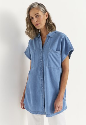 Kobieta o szarych włosach do ramion, ubrana w luźną niebieską bezrękawnikową koszulę na guziki z naszywanymi kieszeniami, stojąca na białym tle.