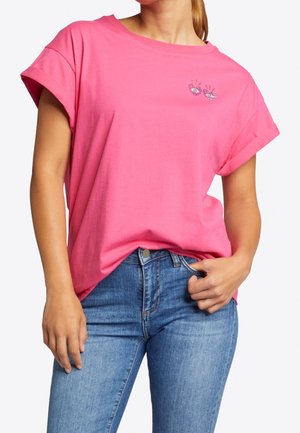 Rosa Baumwoll-T-Shirt mit kurzen, aufgerollten Ärmeln, versehen mit einem kleinen gestickten Motiv auf der Brust. Kombiniert mit blauen, eng anliegenden Jeans.