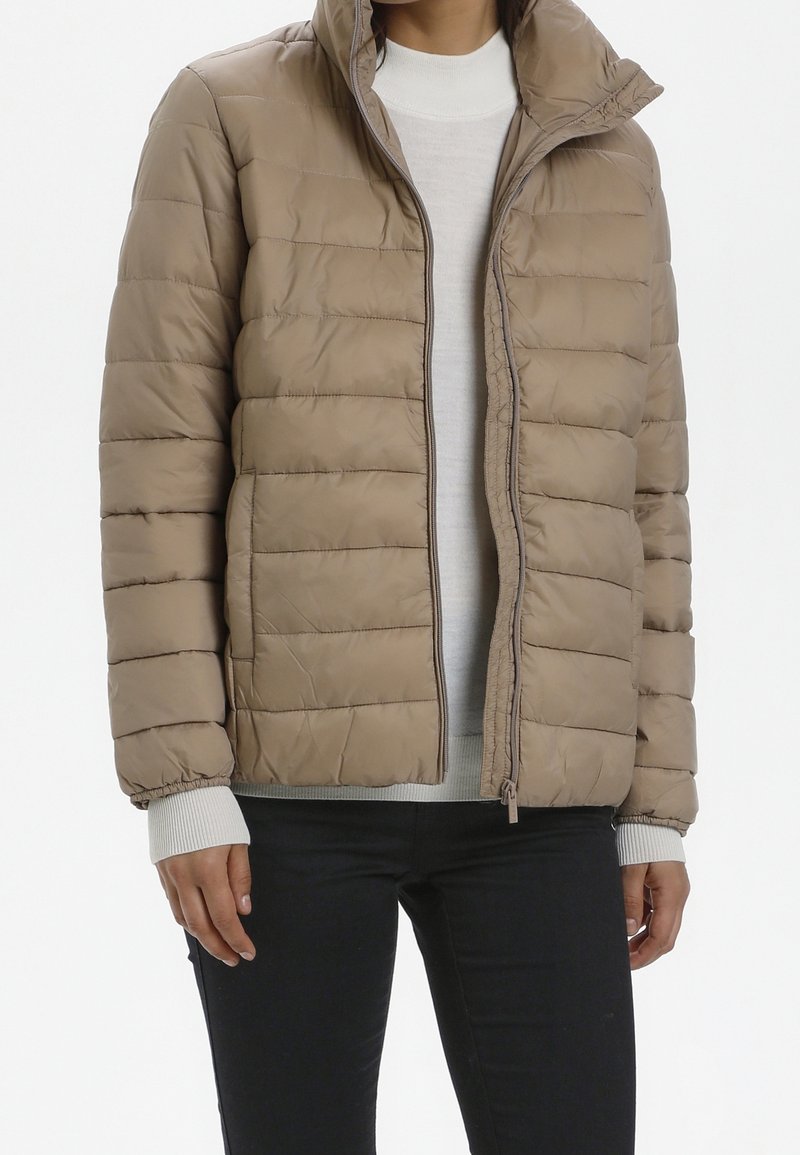 Femme portant une veste matelassée beige par-dessus un pull blanc et un pantalon noir, debout les mains détendues le long du corps.