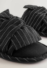 Black slippers with a textured fabric featuring white vertical stripes. The design includes a knotted upper and a soft, padded sole.