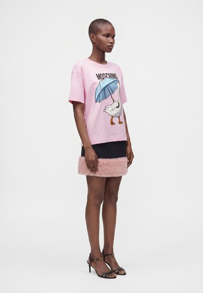 Γυναίκα με κοντά μαλλιά φοράει ένα ροζ t-shirt Moschino με στάμπα πάπιας και ομπρέλας, μια μαύρη μίνι φούστα με ροζ γούνινη επένδυση και μαύρες γόβες.
